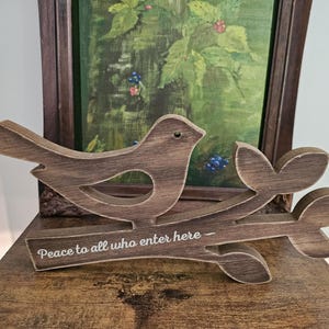 May include: A brown wooden cutout of a bird perched on a branch with leaves. The branch features the white printed text, Peace to all who enter here. The wood has a distressed finish with visible grain patterns and a dark brown stained appearance.