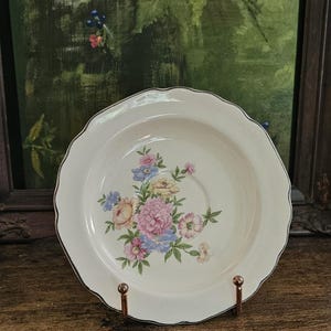 May include: A round, off-white ceramic saucer with a scalloped edge and a silver-toned rim. The center of the saucer features a floral pattern with pink, yellow, and blue flowers surrounded by green leaves and stems on a smooth, glossy surface.