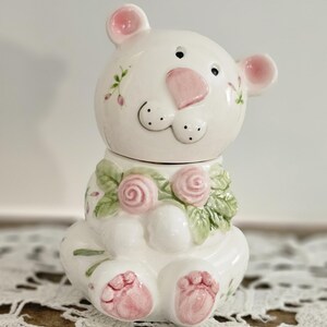 May include: A white ceramic bear figurine featuring a glossy finish, pink painted nose, ears, and paw pads. The bear is adorned with a sculpted collar of green leaves and pink roses, with additional delicate floral accents painted on its body.