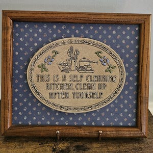 May include: A rectangular wooden frame containing a blue fabric background with a small floral pattern. Centered is an oval plaque with a beige textured surface featuring illustrations of kitchenware and the text: THIS IS A SELF CLEANING KITCHEN, CLEAN UP AFTER YOURSELF.