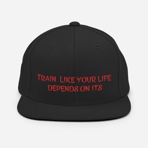 Könnte beinhalten: Schwarze Baseballkappe mit flachem Schirm und dem rot gestickten Text "TRAIN LIKE YOUR LIFE DEPENDS ON ITS". Die Kappe hat eine strukturierte Passform und eine runde Krone, passend für verschiedene Kopfgrößen.