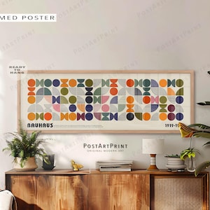 Op de afbeelding: Een houten poster met een geometrisch Bauhaus-ontwerp in oranje, blauw, groen en roze. De tekst "BAUHAUS 1919-19" is zichtbaar op de poster. De poster hangt boven een houten kast. Boven staat "WOOD FRAMED POSTER".