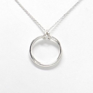 Puede incluir: Un collar plateado con una fina cadena y un colgante circular. El colgante es un diseño de círculo abierto simple, suspendido de la cadena. El collar se muestra sobre una superficie blanca, destacando su estilo minimalista.