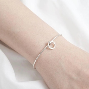 Puede incluir: Una pulsera rígida de tono plateado que presenta una banda de metal fina y lisa que termina en un pequeño elemento de anillo circular. El diseño es sencillo y pulido, mostrando un acabado metálico continuo alrededor de la muñeca.