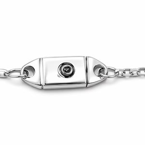 May include: A close-up of a silver-colored bracelet clasp. The clasp is rectangular with rounded ends and a small, dark center stone. The bracelet chain is made of small, circular links.