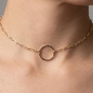 May include: A gold-toned choker necklace featuring a delicate chain with elongated oval links and a central circular pendant. The circular pendant has a textured, hammered surface finish that catches the light, creating a subtle shimmer.