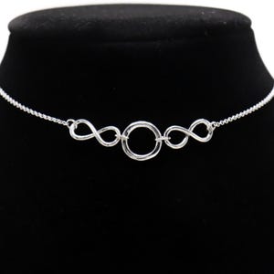 May include: A silver-colored necklace featuring a central circle flanked by two infinity symbols. The necklace is displayed on a black mannequin, showcasing its delicate chain and intricate design. The jewelry is simple and elegant.
