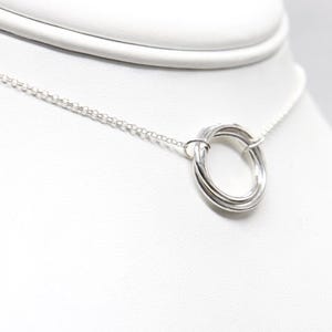 May include: A silver necklace with a delicate chain and a pendant featuring a looped design. The pendant is composed of multiple intertwined bands, creating a textured, circular shape. The necklace is displayed on a white surface.