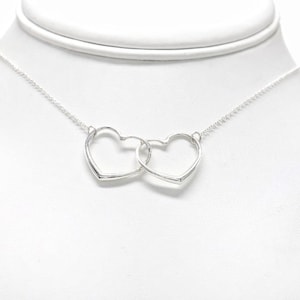Puede incluir: Un collar plateado con dos colgantes entrelazados en forma de corazón. Los corazones, con marco abierto, están conectados y cuelgan de una fina cadena. El collar se muestra sobre un fondo blanco, resaltando el metal brillante y reflectante.