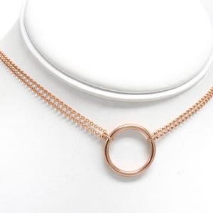 以下が含まれることがあります： ローズゴールドカラーのネックレス。ダブルチェーンのデザインで、円形のペンダントが付いています。白いマネキンのバストに飾られており、繊細なチェーンとミニマルなスタイルが特徴です。このジュエリーはおそらく金属製です。