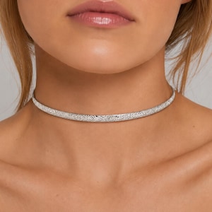 Op de afbeelding: Een zilveren choker ketting met een ingewikkeld bloemmotief. De ketting is een stijve, gebogen band die dicht op de hals zit. Het ontwerp is gedetailleerd en lijkt te zijn geëmbosseerd of gegraveerd.