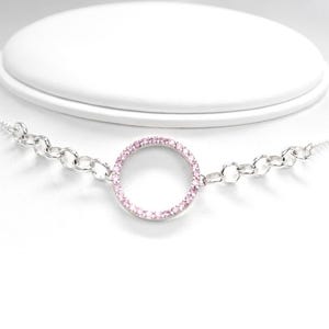 May include: A silver necklace with a circular pendant. The pendant is encrusted with small pink gemstones. The necklace has a chain design on either side of the pendant and is displayed on a white mannequin.
