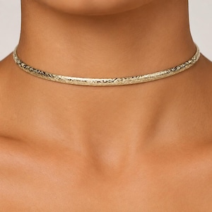 Puede incluir: Un collar tipo gargantilla de metal en tono dorado con una banda delgada y redondeada que presenta una superficie texturizada y martillada. La pieza se ajusta al cuello, mostrando un brillo metálico constante y un patrón detallado e irregular.