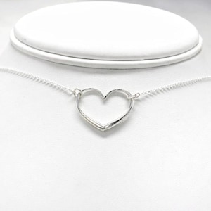 May include: A silver-colored necklace featuring a heart-shaped pendant. The pendant is a simple, open heart design suspended from a delicate chain. The necklace is displayed on a white bust, showcasing its elegant and minimalist style. A perfect gift for a loved one.
