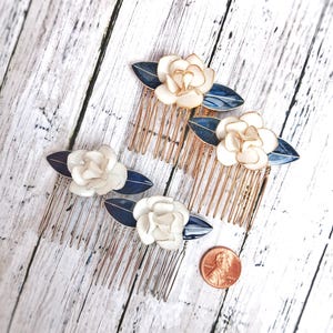 May include: Four hair combs with white flower and navy blue leaf accents. The combs have gold and silver metal teeth. A penny is next to the combs for size reference. The combs are on a white and gray wooden surface.