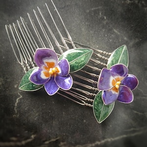 May include: Two silver hair combs with purple and white flower designs and green leaves. The flowers have yellow centers. The combs are set against a dark background.
