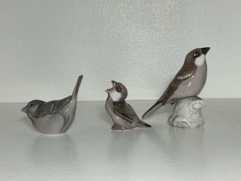 May include: Three ceramic bird figurines in shades of gray and brown. One bird is perched on a small, textured base. Another is in a singing pose, and the third is in a resting position.