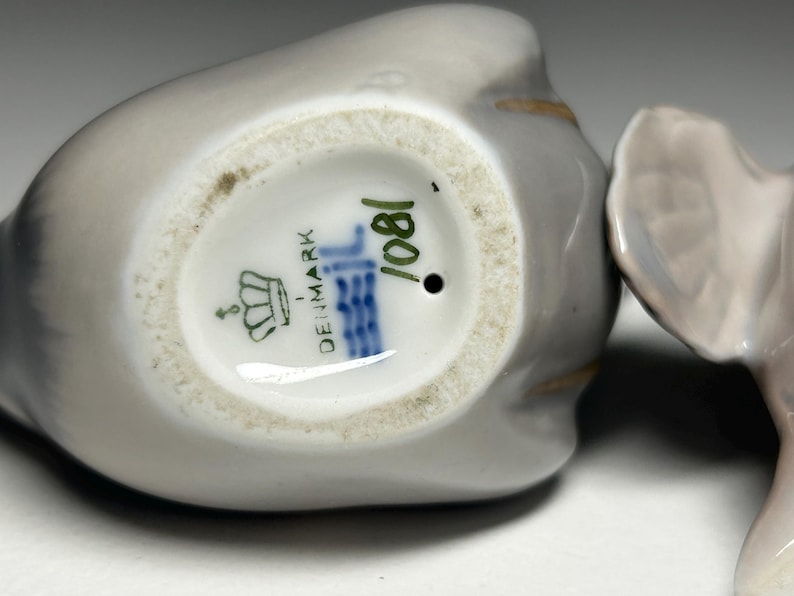 May include: Close-up of a porcelain figurine, likely a bird, with a gray and white color scheme. The base of the figurine is visible, displaying the text "DENMARK" and the number "1081". The figurine has a glossy finish.