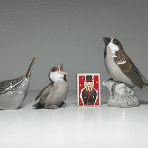 May include: Three ceramic bird figurines in shades of gray and brown, with a matchbox featuring a magician. The birds vary in pose, with one perched, one singing, and one with its head down. A decorative collection.