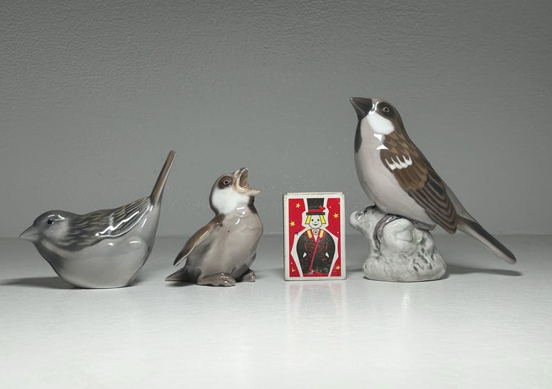 May include: Three ceramic bird figurines in shades of gray and brown, with a matchbox featuring a magician. The birds vary in pose, with one perched, one singing, and one with its head down. A decorative collection.