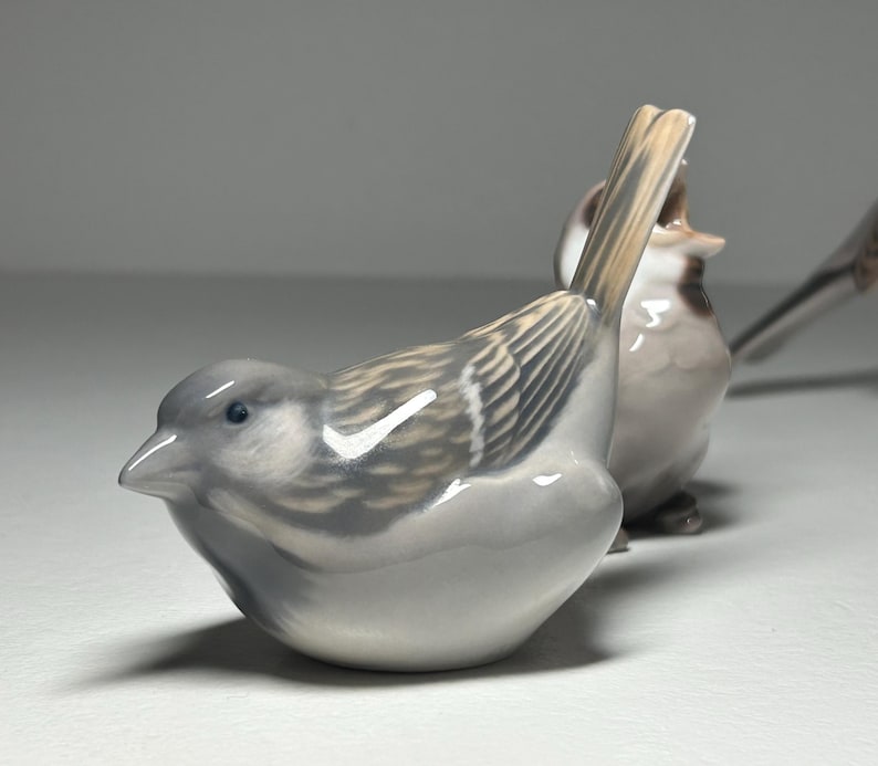 May include: Two ceramic bird figurines. The first bird is in focus, with a gray body, detailed wing patterns, and a light brown tail. The second bird is slightly out of focus, with a similar design. The figurines are on a white surface.