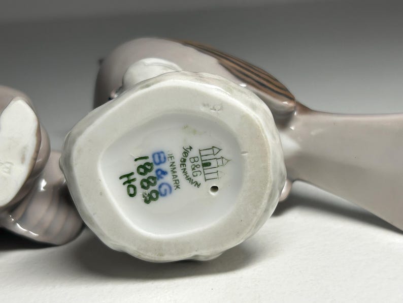 May include: A pair of ceramic bird figurines in shades of gray and brown. The underside of one bird shows the B&G Copenhagen Denmark mark, along with the numbers 1883 and HO in green ink. The birds are likely decorative items.