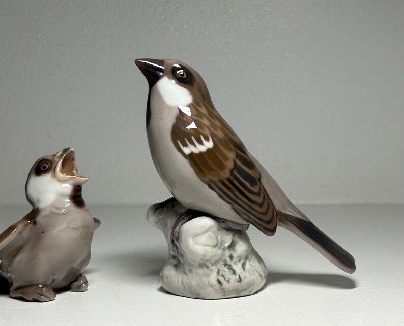 May include: Two ceramic bird figurines. One is a sparrow perched on a rock, with brown, white, and gray feathers. The other is a smaller bird with an open beak, in shades of brown and white. The figurines are on a white surface.