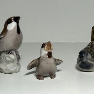 May include: Three ceramic bird figurines. One bird is perched on a rock, another has its wings spread, and the third is in a resting position. The birds are primarily gray, brown, and white.