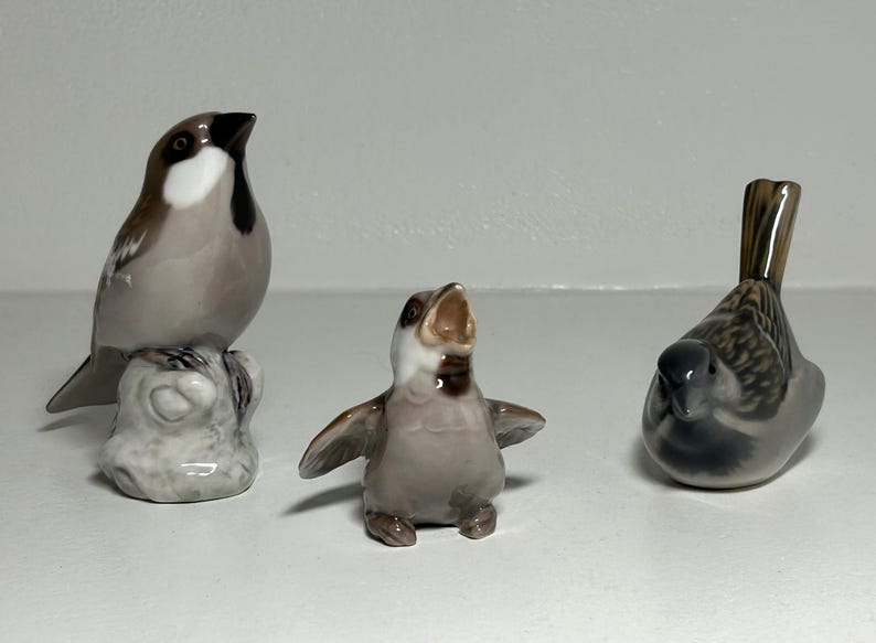 May include: Three ceramic bird figurines. One bird is perched on a rock, another has its wings spread, and the third is in a resting position. The birds are primarily gray, brown, and white.