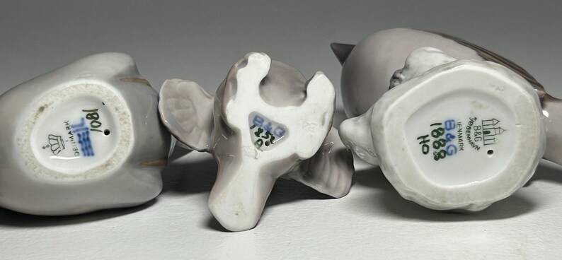 May include: Three ceramic bird figurines in shades of gray, with visible markings on the underside. The figurines are in various poses, showcasing their detailed craftsmanship. The bases display maker's marks and numbers, indicating their origin and design.