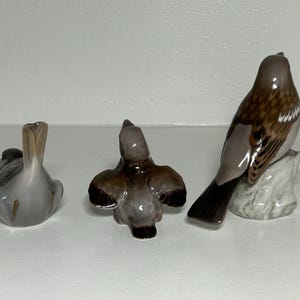 May include: Three ceramic bird figurines in various poses. One bird is sitting, another is in flight, and the third is perched on a small rock. The birds have brown, gray, and white coloring.