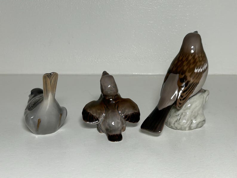 May include: Three ceramic bird figurines in various poses. One bird is sitting, another is in flight, and the third is perched on a small rock. The birds have brown, gray, and white coloring.