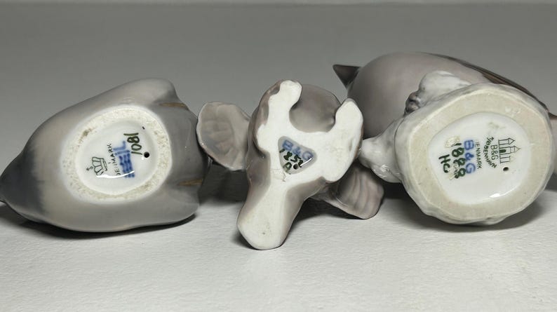 May include: Three gray ceramic bird figurines with visible markings on the bottom. The figurines are in various poses, with one lying on its side. The markings include text and a crown symbol.