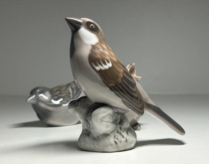 May include: Two ceramic bird figurines. One bird is perched on a gray rock, with brown and white feathers. The other bird is in the background, in a resting position. The figurines are detailed and have a glossy finish.