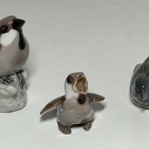 May include: Three ceramic bird figurines. One is perched on a rock, another has its beak open, and the third is facing down. The birds are primarily gray, brown, and white, with detailed feather patterns. A decorative set.