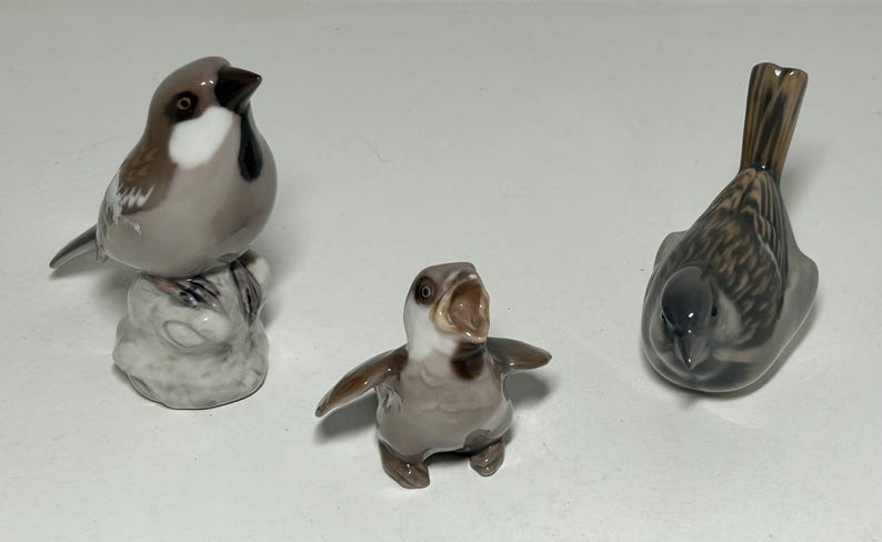 May include: Three ceramic bird figurines. One is perched on a rock, another has its beak open, and the third is facing down. The birds are primarily gray, brown, and white, with detailed feather patterns. A decorative set.