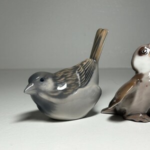 May include: Two ceramic bird figurines. One is a gray sparrow with brown and white wing details, perched with its tail up. The other is a brown and gray bird with its beak open, as if singing. These decorative figurines are ideal for home decor.