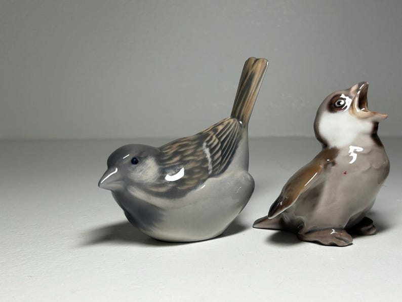 May include: Two ceramic bird figurines. One is a gray sparrow with brown and white wing details, perched with its tail up. The other is a brown and gray bird with its beak open, as if singing. These decorative figurines are ideal for home decor.