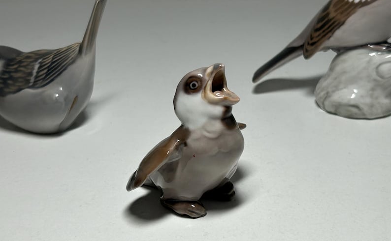 May include: Three ceramic bird figurines in shades of gray, brown, and white. One bird is depicted with its beak open, as if singing. The other two birds are in different poses. The figurines are on a white surface.