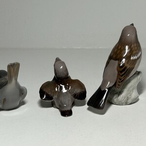 May include: Three ceramic bird figurines in varying poses. The birds are primarily gray, brown, and white, with detailed feather patterns. One bird is perched on a small, textured base. The figurines are displayed on a white surface.