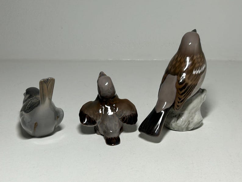 May include: Three ceramic bird figurines in varying poses. The birds are primarily gray, brown, and white, with detailed feather patterns. One bird is perched on a small, textured base. The figurines are displayed on a white surface.