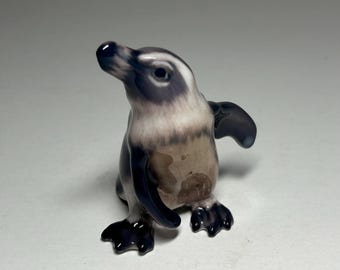 May include: A small ceramic penguin figurine with black and white markings. The penguin is standing on a white surface.