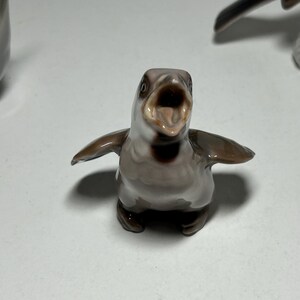 May include: A ceramic penguin figurine with an open beak and outstretched wings. The penguin is primarily gray and brown with a white belly. The figurine is a decorative item.