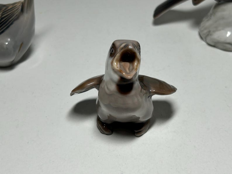 May include: A ceramic penguin figurine with an open beak and outstretched wings. The penguin is primarily gray and brown with a white belly. The figurine is a decorative item.