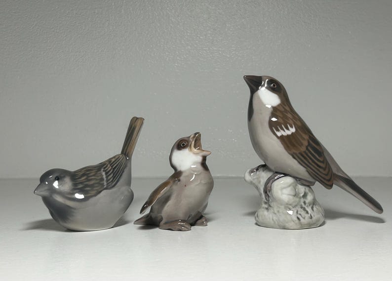May include: Three ceramic bird figurines in shades of gray, brown, and white. One bird is perched on a rock, another has its beak open, and the third is in a resting position. The figurines are detailed and realistic.