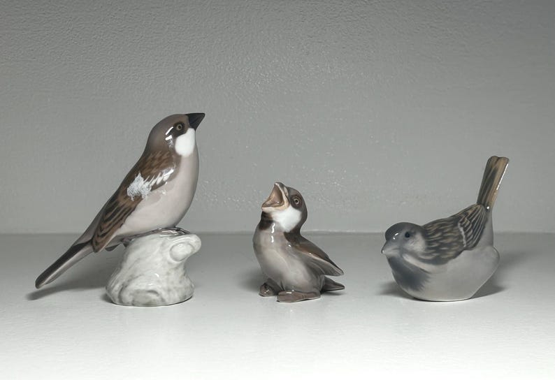 May include: Three ceramic bird figurines in shades of gray, brown, and white. One bird is perched on a textured base, another has its beak open, and the third is in a resting position. The figurines are detailed and appear to be decorative.