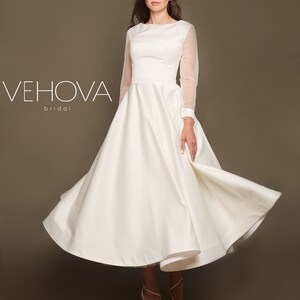 Puede incluir: Un vestido de novia de sat&eacute;n blanco con una falda completa y mangas largas transparentes. El vestido tiene un dise&ntilde;o simple y elegante, perfecto para una boda cl&aacute;sica.