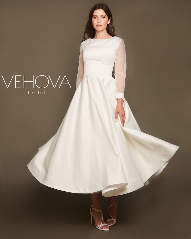 Puede incluir: Un vestido de novia de sat&eacute;n blanco con una falda completa y mangas largas transparentes. El vestido tiene un dise&ntilde;o simple y elegante, perfecto para una boda cl&aacute;sica.