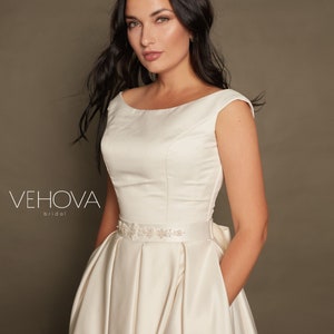 Peut inclure: Une robe de mari&eacute;e en satin blanc avec un d&eacute;collet&eacute; en bateau et des manches courtes. La robe a un corsage ajust&eacute; et une jupe ample. Une ceinture perl&eacute;e cintr&eacute;e la taille.