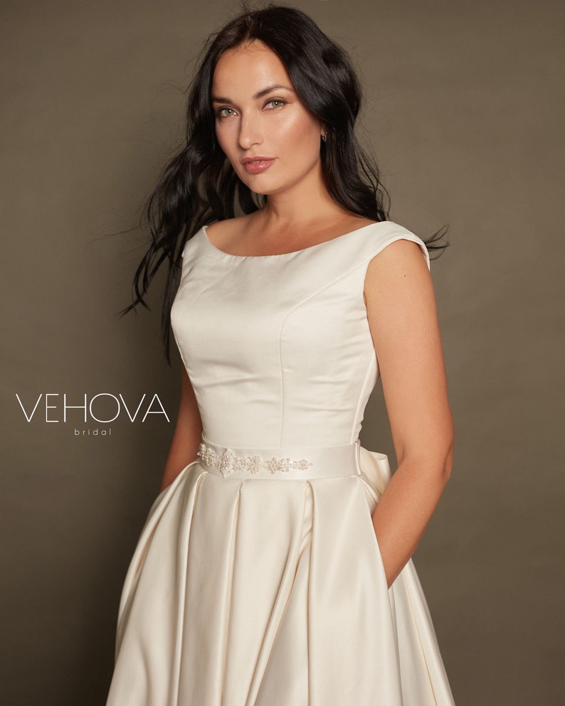 Peut inclure: Une robe de mari&eacute;e en satin blanc avec un d&eacute;collet&eacute; en bateau et des manches courtes. La robe a un corsage ajust&eacute; et une jupe ample. Une ceinture perl&eacute;e cintr&eacute;e la taille.